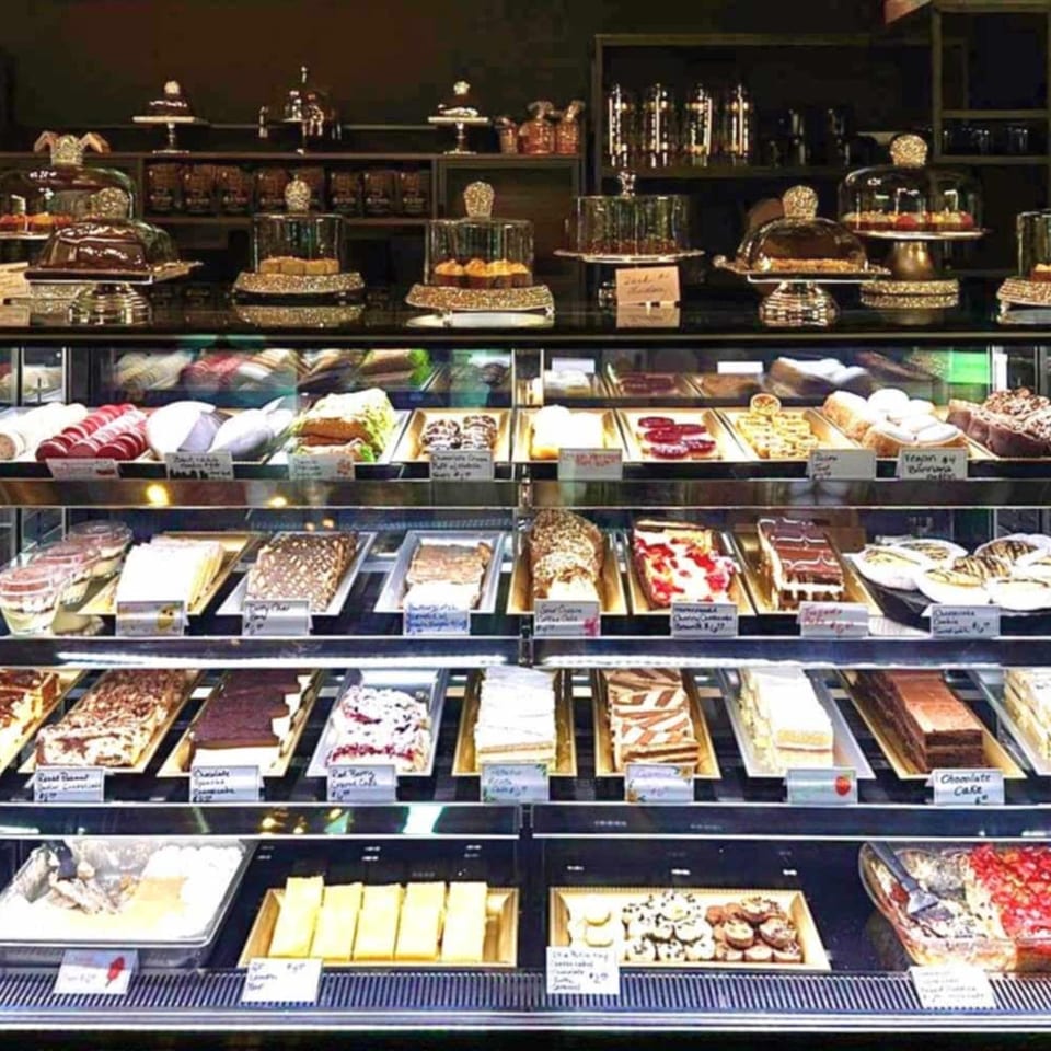 A brightly lit display case filled with various cakes, pastries, and desserts in a bakery.