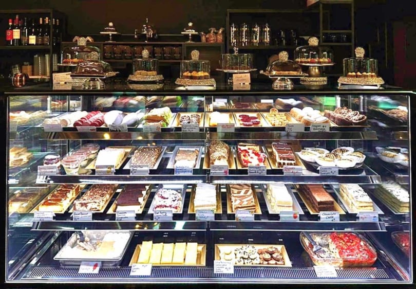 A brightly lit display case filled with various cakes, pastries, and desserts in a bakery.