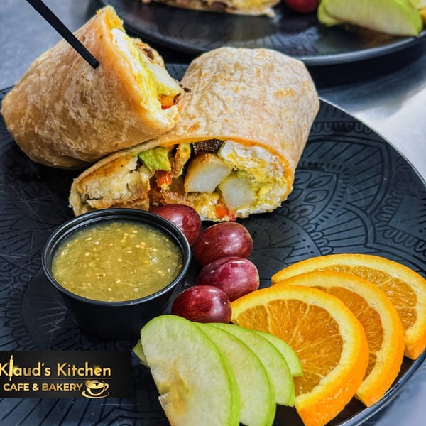 Two breakfast burritos with salsa verde, grapes, orange slices, and green apple slices on black plates.