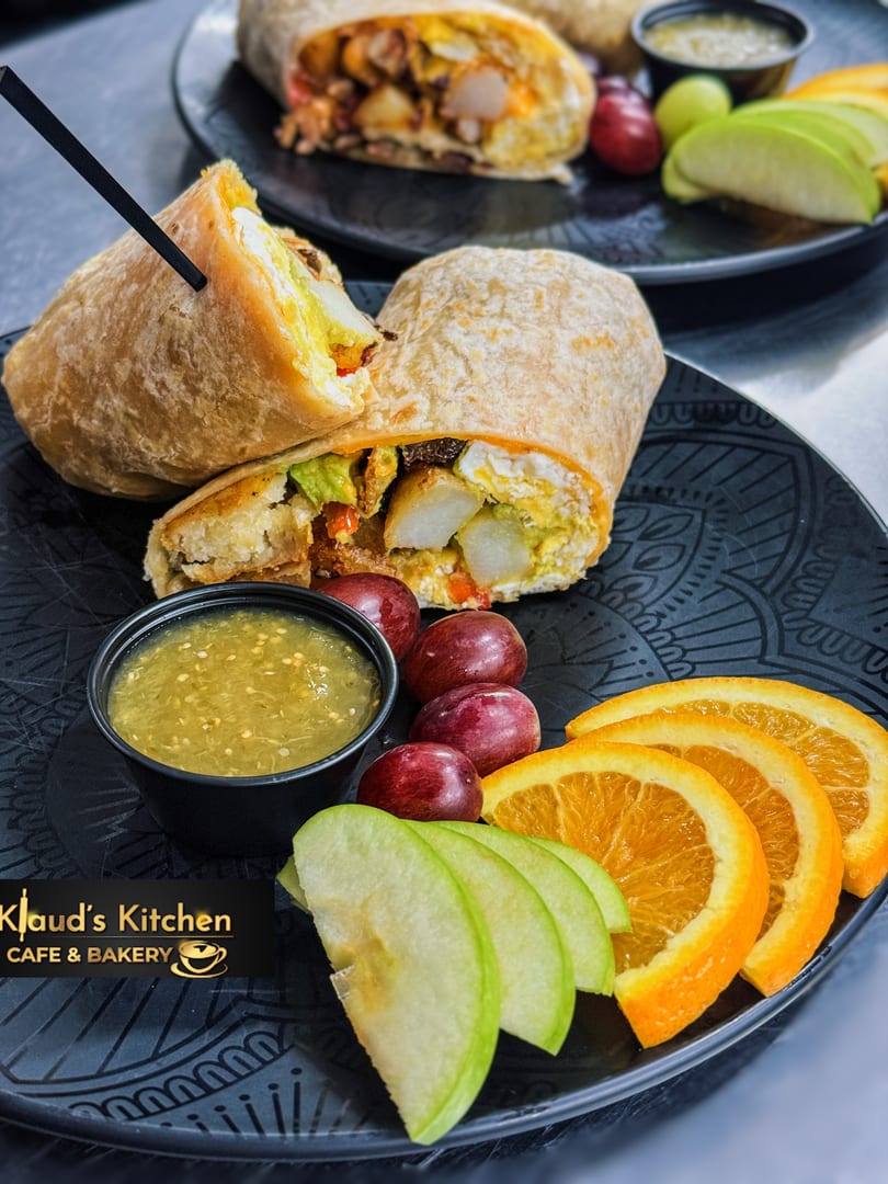 Two breakfast burritos with salsa verde, grapes, orange slices, and green apple slices on black plates.
