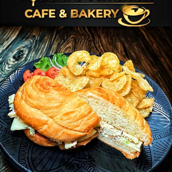 A croissant sandwich with chips, lettuce, tomatoes, and onions on a dark patterned plate.