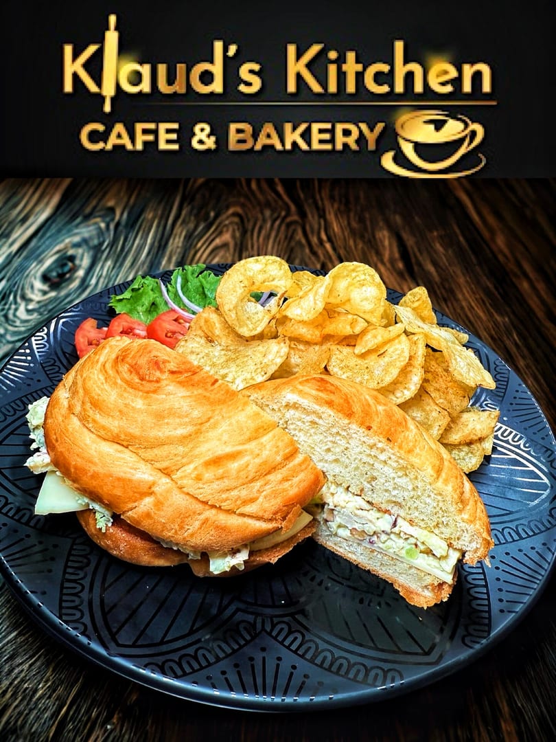 A croissant sandwich with chips, lettuce, tomatoes, and onions on a dark patterned plate.