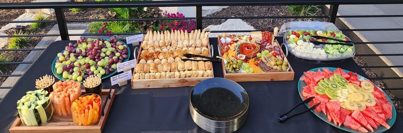 An outdoor buffet table with grapes, pastries, charcuterie, vegetables, and fruit platters.