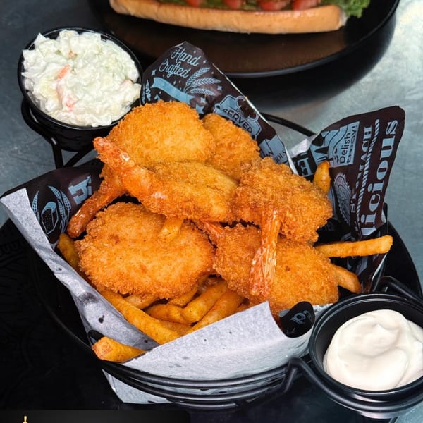 A basket of crispy fried shrimp and fries, with coleslaw, dipping sauce, and a sandwich in the background.