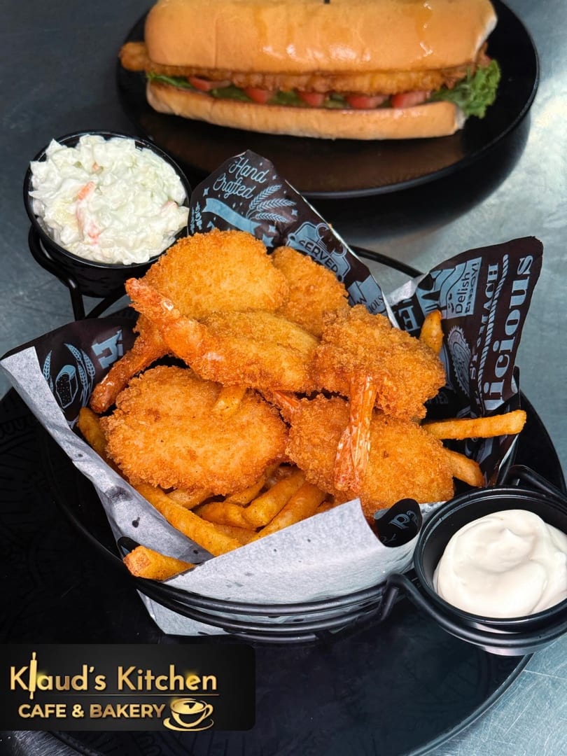 A basket of crispy fried shrimp and fries, with coleslaw, dipping sauce, and a sandwich in the background.